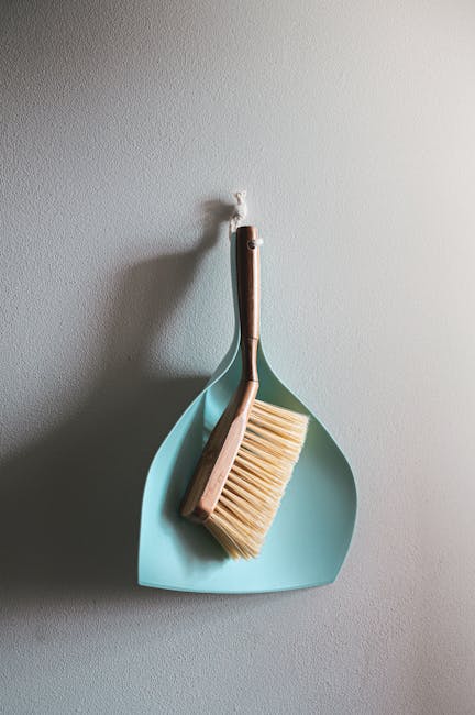 A close-up of a light blue dustpan hanging on a white textured wall, with a wooden-handled broom and a beige scrub brush resting inside it. The surfaces appear clean, with the wooden handle of the bro