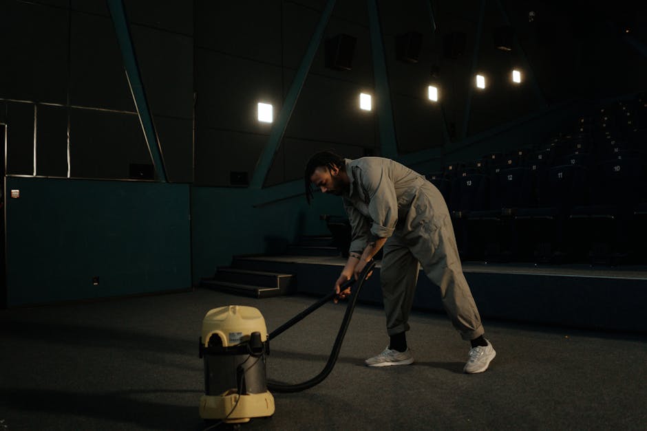 A person is kneeling on a floral patterned carpet, using a yellow and black vacuum cleaner for surface cleaning and deep cleaning of the carpet. The individual is wiping the vacuum's dust compartment 