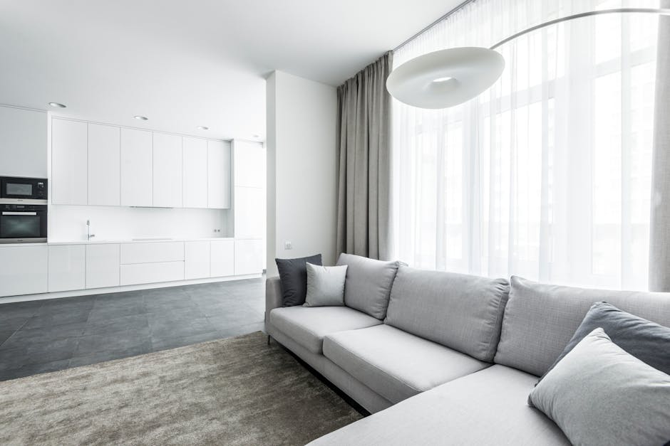 A beige fabric sofa with plush cushions positioned against a white wall with decorative molding, situated on a light grey carpeted floor in a well-lit living room. The room features a classic architec